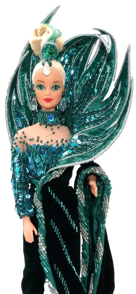 Boneca Barbie loira com maquiagem marcante, vestindo um traje de gala verde-esmeralda e azul-turquesa, com lantejoulas, mi&ccedil;angas e detalhes prateados. Ela usa um adere&ccedil;o de cabe&ccedil;a e asas grandes, que se assemelham a penas, adornadas com brilhos. O vestido tem uma gola alta e mangas longas, com um corpete transparente na parte superior. A boneca est&aacute; em p&eacute;, com o bra&ccedil;o direito levemente dobrado, em um fundo branco