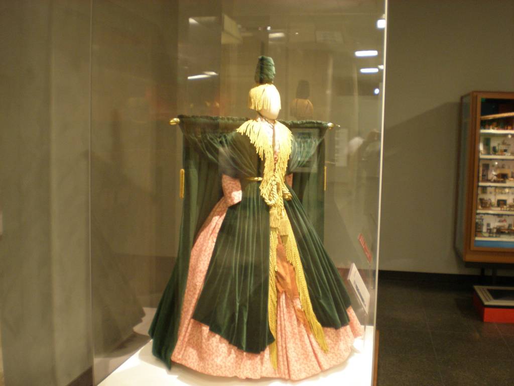 Vestido de baile verde-escuro e rosa, com corpete dourado e franjas, exposto em vitrine de vidro em um museu